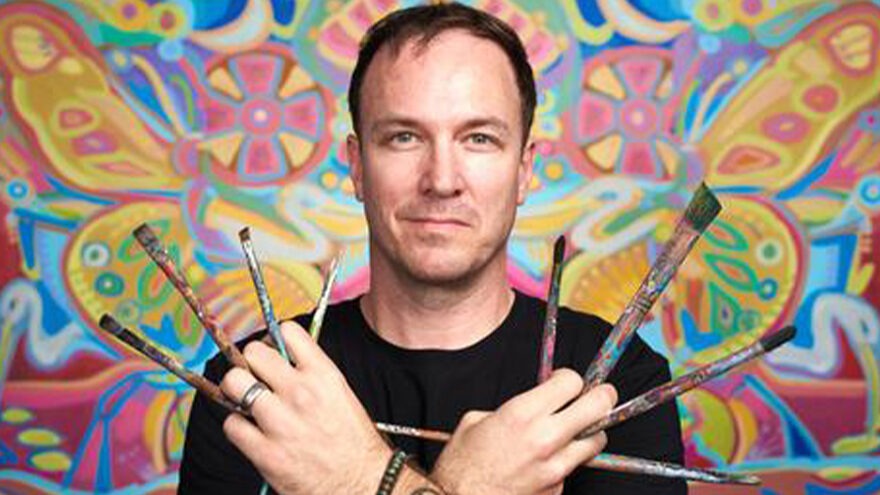 Performance artist and painter John Bukaty posing in front of a mural with paintbrushes between his fingers