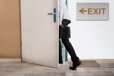 businessperson walking out of a door with exit sign on wall, representing his resignation