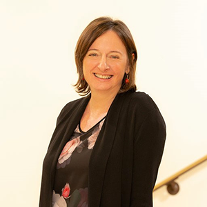A portrait of Sandy hammer. She is a white woman with shoulder-length brown hair and a black cardigan