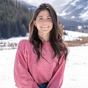 A portrait of Bree DiBernardo. She is a white woman with wavy brown hair and a pink sweater standing in the snow