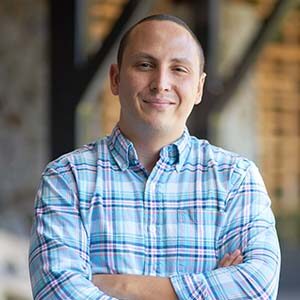 A portrait of Ryan Grande. He is a white man with shaved brown air and a blue plaid collared shirt