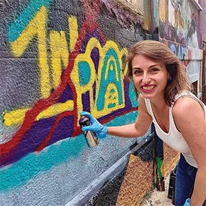 A portrait of Ashley Lawson. She is a white woman with short brown hair and a white tank top. She is holding a purple spray paint can against a graffitied wall
