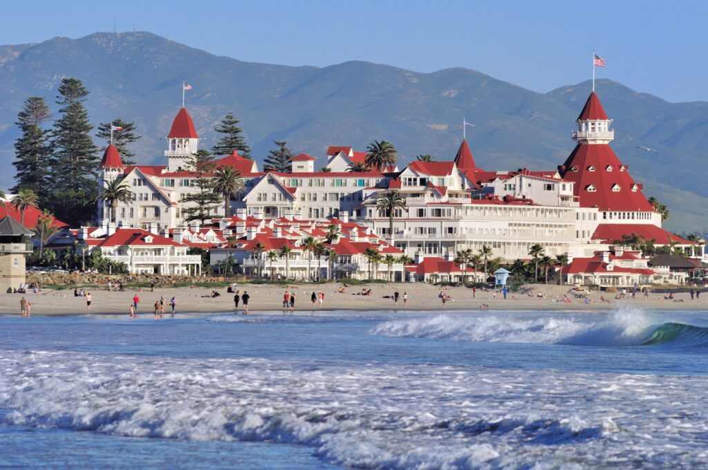 Hotel del Coronado. It is a large white building on the beach with a series of red sloping roofs. Mountains are in the distance 