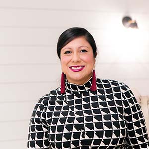 A portrait of Bianca Ferrer. She is a brown woman with red earrings and a black and white patterned blouse