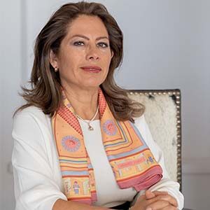 A portrait of Claudia Perez. She is a white woman with wavy brown hair and a pastel patterned ascot