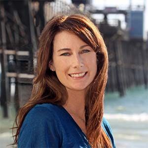 A portrait of Dana Porter Higgins. She is a white woman with wavy brown hair and a blue blouse standing on a beach