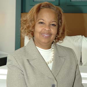 A portrait of Jacquelyn Denise Reid. She is a black woman with curly reddish hair, a pearl necklace and a grey suit jacket