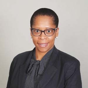 A portrait of Kim Hughes. She is a black woman with hair in a bun, cat eye glasses and a dark grey suit jacket