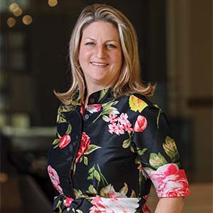 A portrait of Kirsten Olean. She is a white woman with straight blonde hair and a black floral blouse