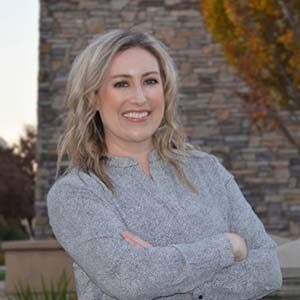 A portrait of Maggie Thyken Huss. She is a white woman with wavy blonde hair and a grey sweater