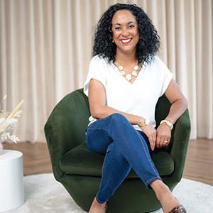 A portrait of Mahoganey Jones. She is a black woman with shoulder-length natural hair and a white blouse. She is sitting in a green armchair