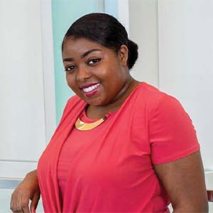 A portrait of Shamera Stewart. She is a black woman with hair in a bun and a pink cardigan