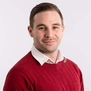 A portrait of Shane Lewis. He is a white man with combed brown hair, a stubbled beard and a red sweater