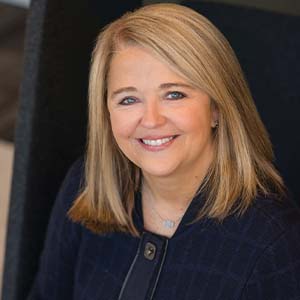 A portrait of Tammy Routh. She is a blonde white woman with straight blonde hair and a dark blue blouse