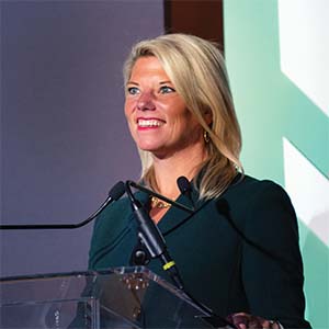 A picture of Tori Emerson Barnes. She is a blonde white woman with a dark blouse. She is speaking at a podium