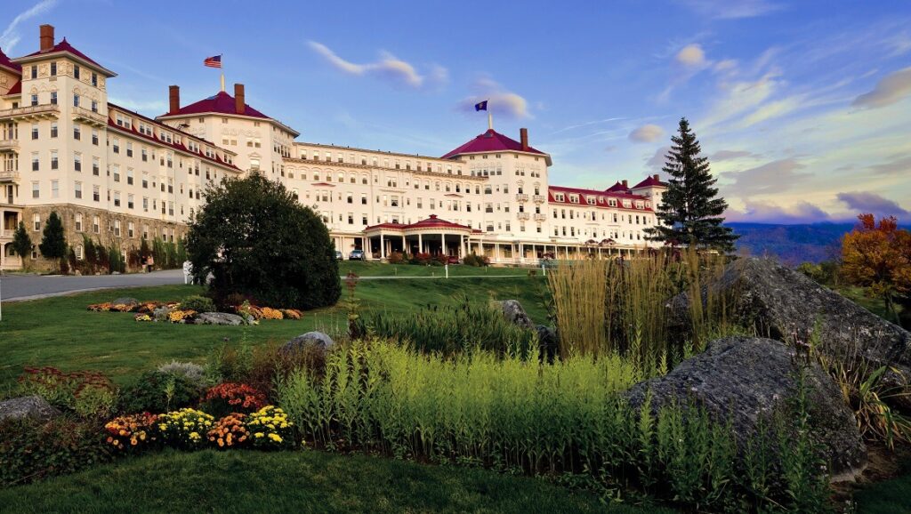 Omni Hotel Mount Washington. It is a white castle-like building with a red roof and angular turrets. A lush garden and lawn are in front, and hills are in the distance