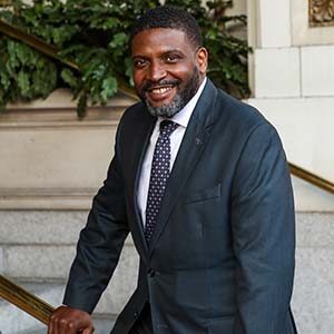 A portrait of Cole Wallace. He is a black man with buzzed hair, a greying beard and a dark suit