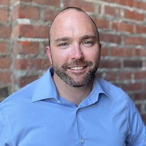 A portrait of Matt Skaletsky. He is a bald man with a stubbled beard and blue collared shirt