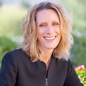 A portrait of Paige Lund. She is a blonde white woman with wavy shoulder-length hair and a black jacket