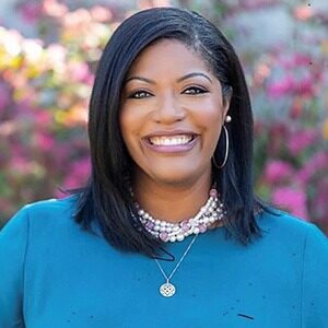 A portrait of Tenille Perry. She is a black woman with shoulder-length straight hair, white necklaces and a blue blouse