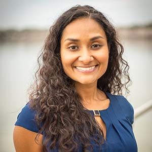 A portrait of Sophia Hyder Hock. She is a South Asian woman with curly brown hair and a blue blouse