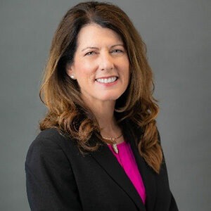 A portrait of Susan Valen. She is a white woman with long wavy brown hair and a black suit