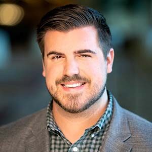 A portrait of Tanner Austin. He is a white man with combed black hair, a stubbled beard and grey suit