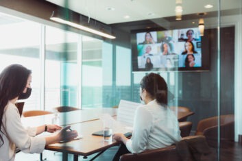Two business women at a conference table with a screen displaying other colleagues' faces