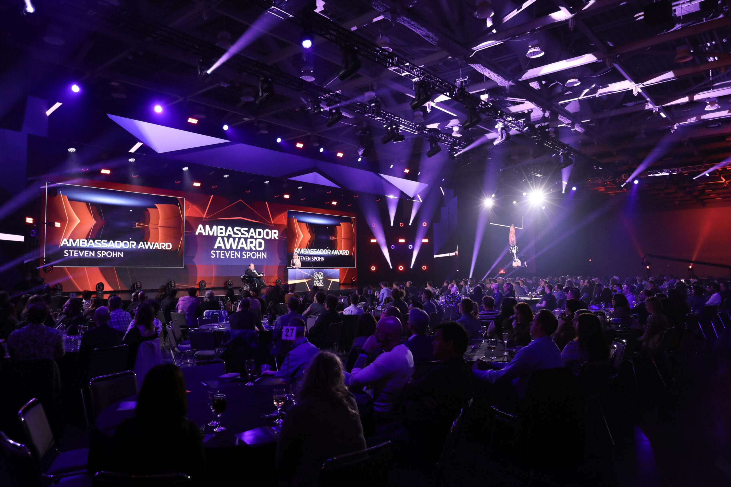 An announcer onstage at the 24th annual Independent Games Festival Awards in California. A crowd sitting at round tables watches.