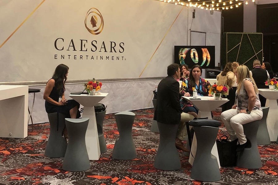 Event attendees sit at small square tables with a modernist design. A Caesars Entertainment banner is behind them.