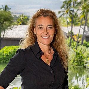 A portrait of Sandra Estornell. She is a tan woman with curly blonde hair and a black collared shirt.