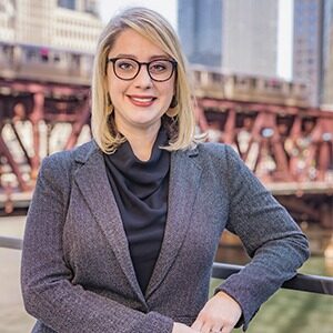 A portrait of Dina Fenili Niekamp. She is a white woman with round glasses, straight blonde hair and a gray suit jacket.