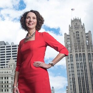A portrait of Lynn Osmond. She is a white woman with short curly hair in a red dress. She stands in front of Chicago skyscrapers.
