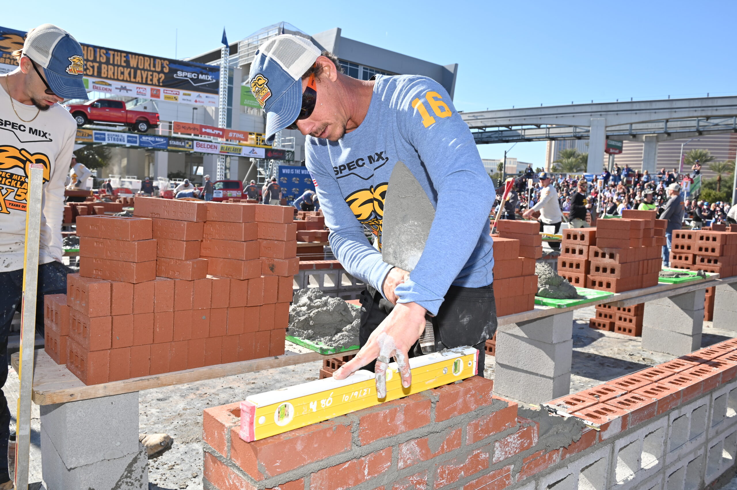 Two men laying bricks by hand in a bricklaying competition. A crowd and car advertisement are in the background.