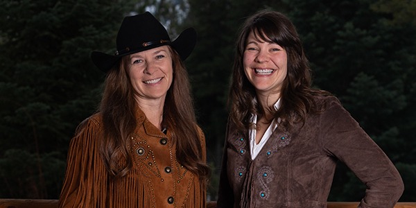 A portrait of Kristen Snavely and Molly Smith. They are both white women with long brown hair wearing cowboy outfits.