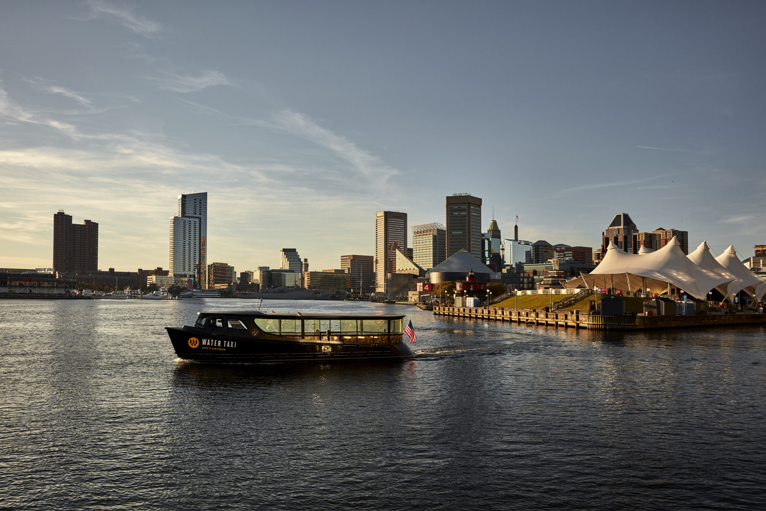 A picture of the Warner Street District in Baltimore. A water taxi is driving by a grassy hill covered in tents, with skyscrapers in the background.