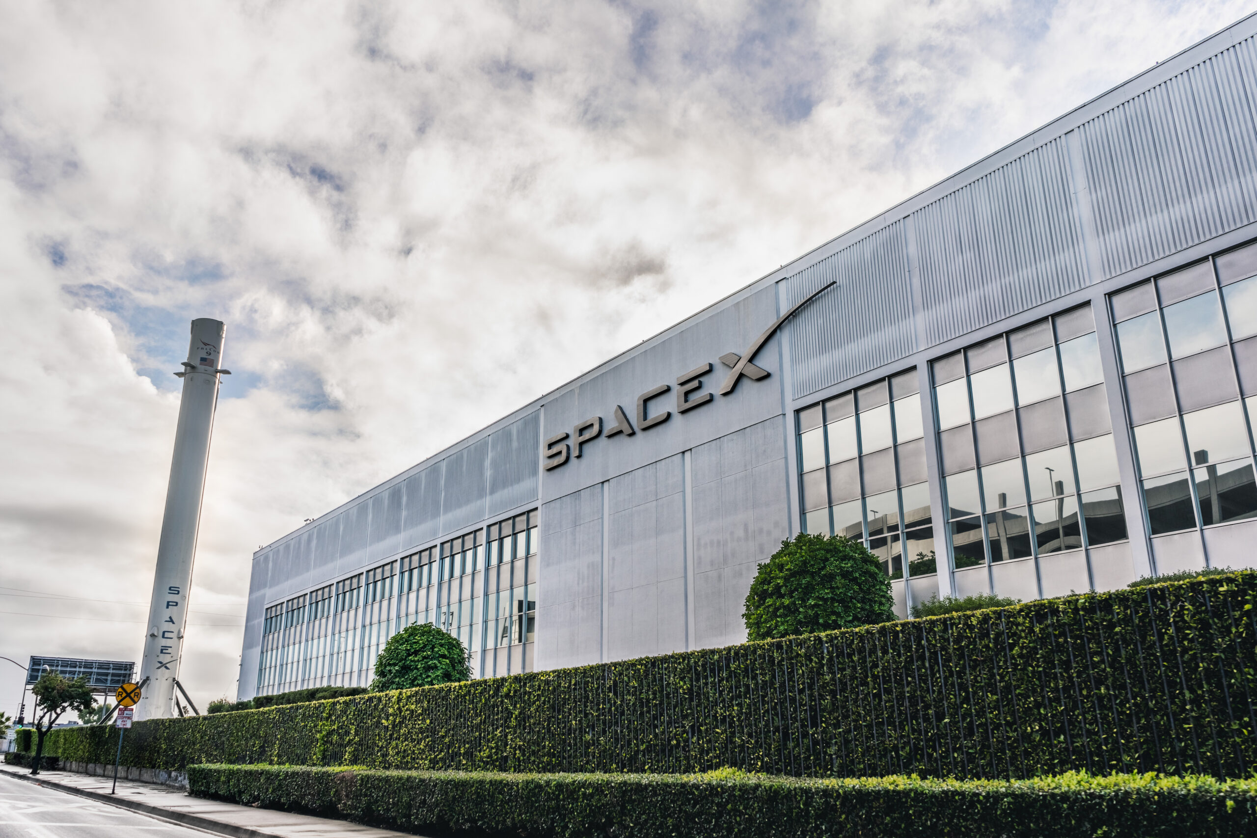 The outside of SpaceX headquarters in Hawthorne, California. It is a grey warehouse surrounded by a short hedge