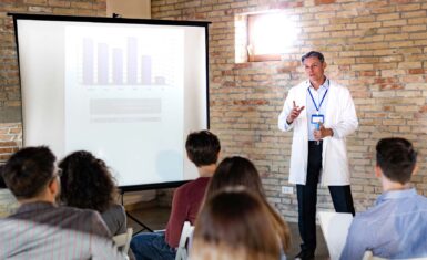 A person in a lab coat explains the data on a projector to a small group of people