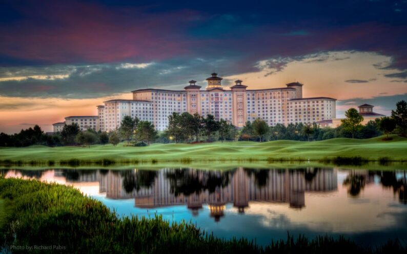 An exterior shot of Rosen Shingle Creek, a property in Orlando. It sits behind a reflective lake on a grassy hill.