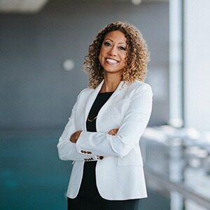 A portrait of Angela Val. She is a black woman wearing a white suit jacket and black dress.