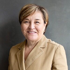 A portrait of Anna Olson. She is an older white woman with a brown pixie cut and a tan suit jacket.