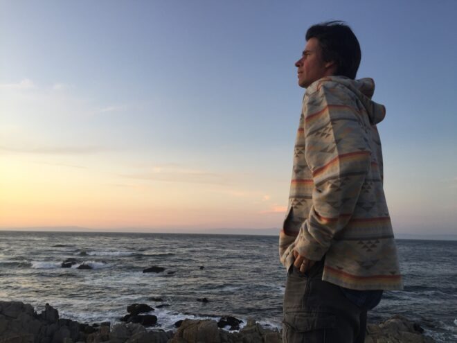 MeetGreen Director of Sustainability Eric Wallinger stands on a rocky beach at sunset with an oversized jacket.
