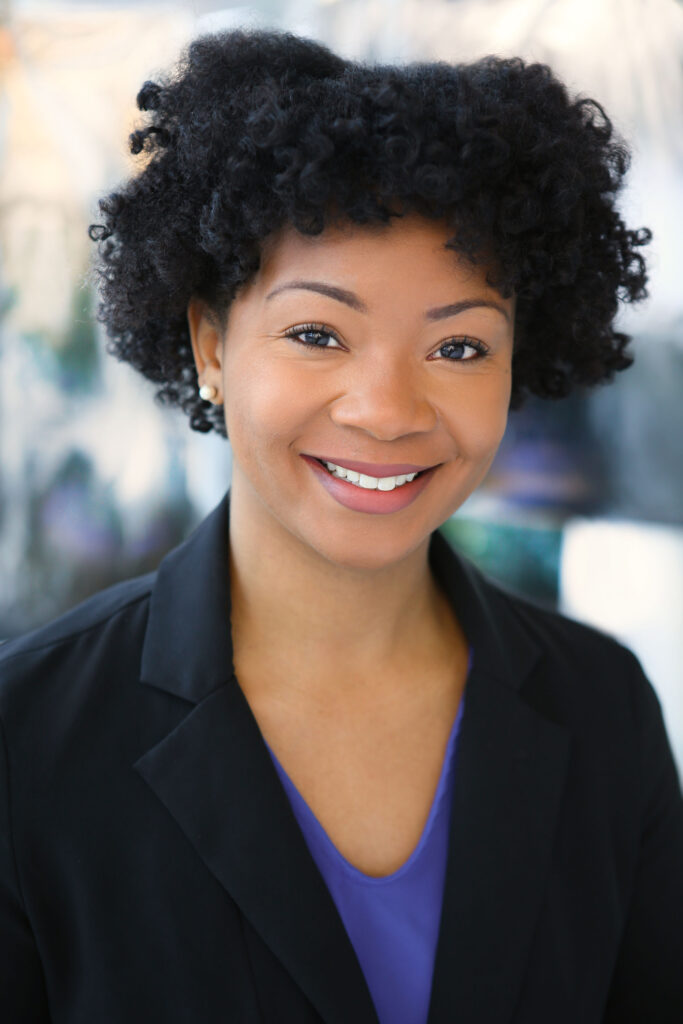 A portrait of Kayla Adams, a young black woman with a short afro and a black suit jacket.