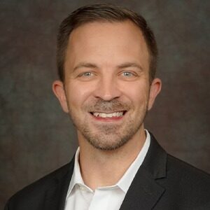 A portrait of Mike Powers. He is a white man with short combed hair, a short beard and a black suit jacket.