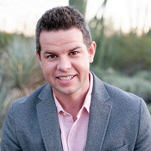 A portrait of Tim Keough. He is a white man with spiked brown hair, a gray suit jacket and a pink collared shirt.