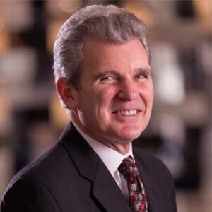 A portrait of Tony Mira. He is an older white man with a black suit and red tie.