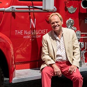 A portrait of Urmas Karner, the new general manager of The Nantucket Hotel & Resort in Massachusetts. He is a blond white man with a tan jacket sitting on the step of a firetruck.