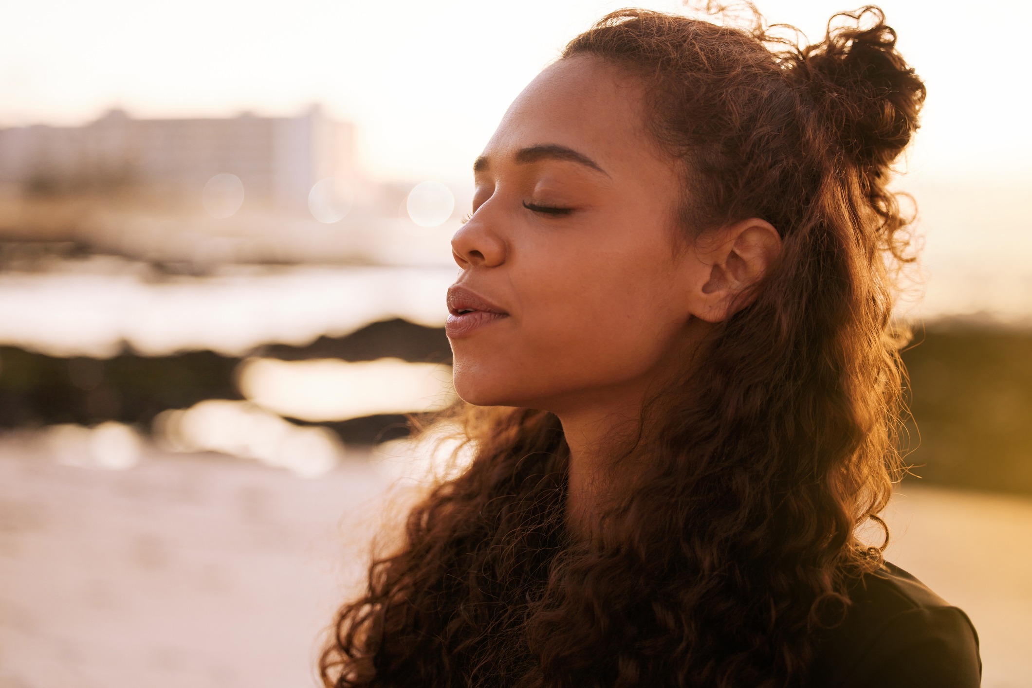 A black woman with loosely curled hair has her eyes closed and is exhaling through her mouth. It is sunset and the background is blurred.