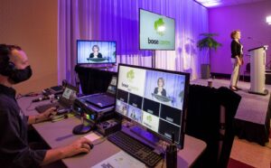 A man wearing a mask and headphones is at an AV booth next to a stage where a woman is giving a presentation.