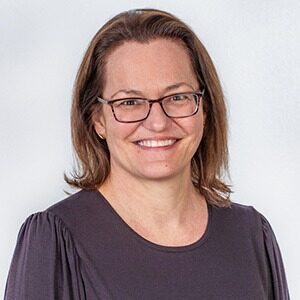 A portrait of Elizabeth Connor. She is a white woman with square glasses and a dark grey blouse.